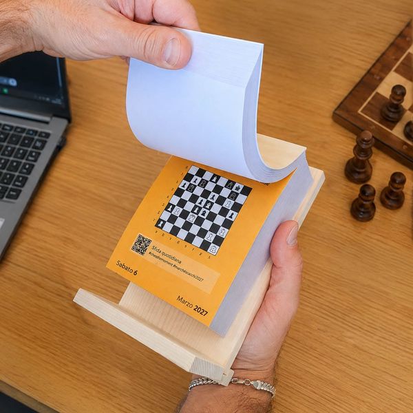 Una mano sfoglia il Calendario degli Scacchi accanto a una scacchiera